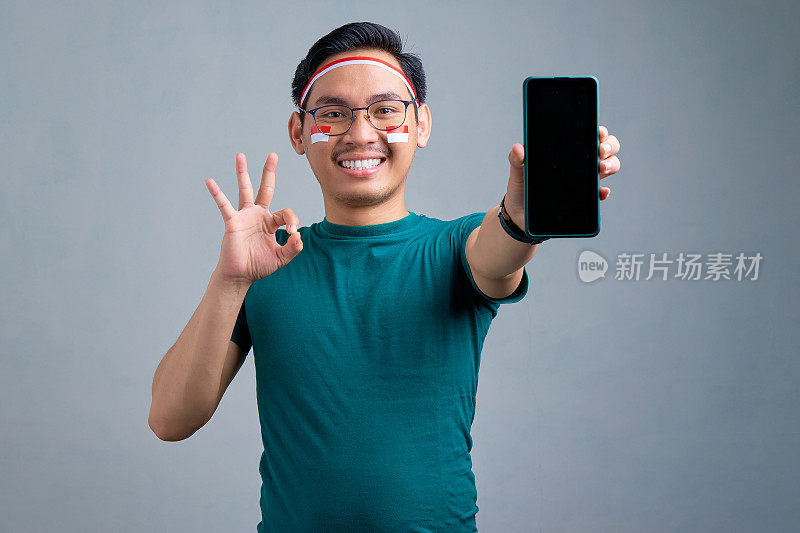 微笑的亚洲年轻男子在休闲t恤显示空白屏幕的手机和手势okay sign孤立的灰色背景。印度尼西亚独立日庆祝概念